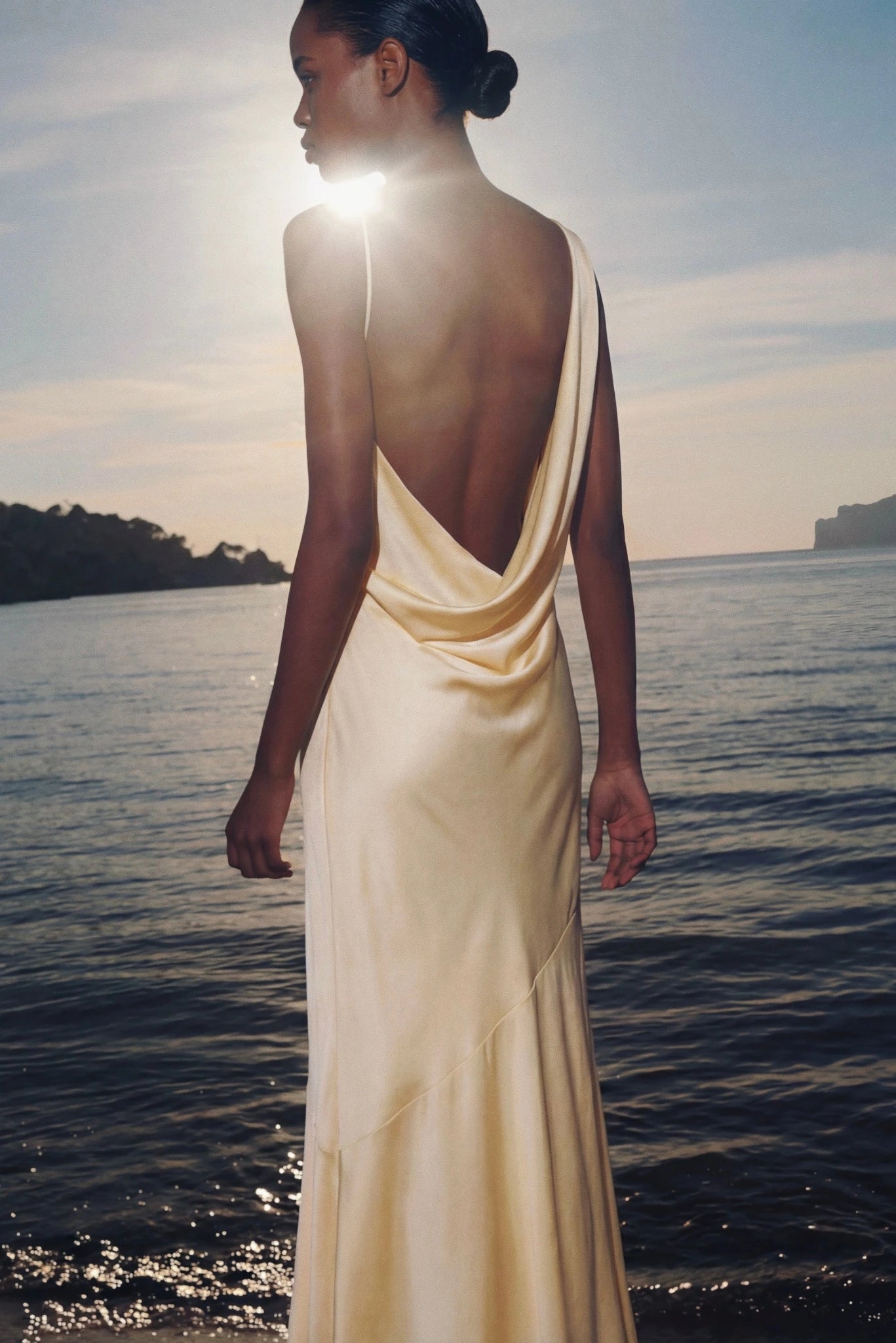 Costas de vestido longo amarelo claro com decote profundo em cenário à beira-mar