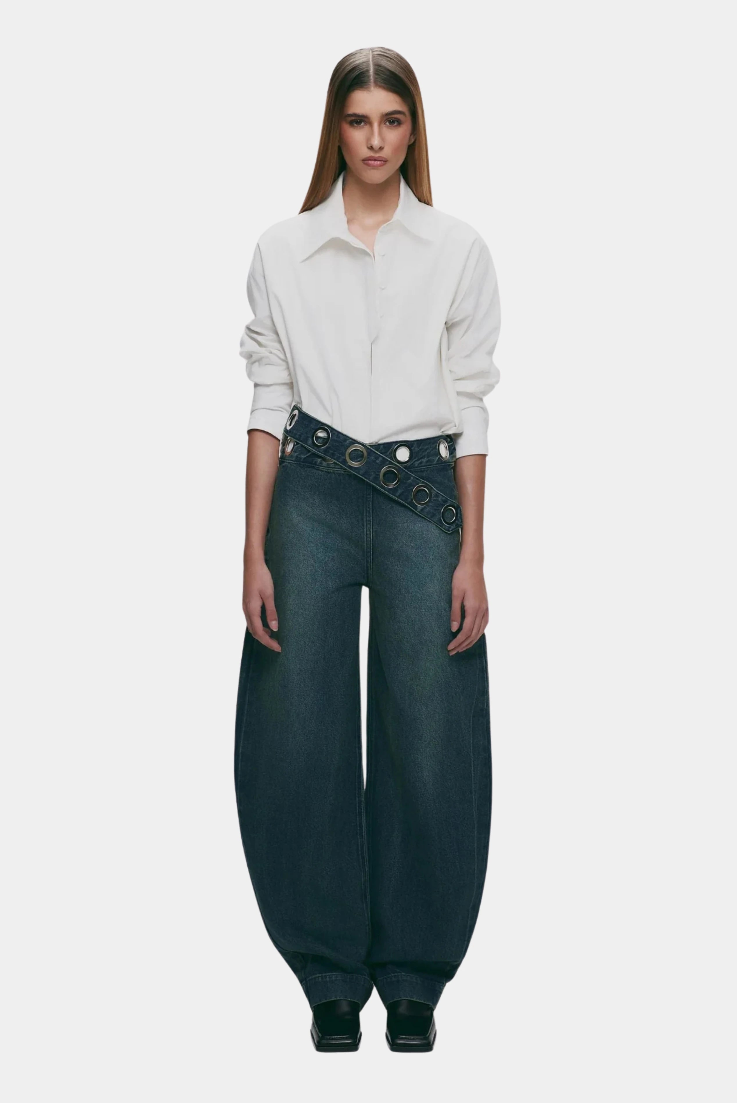 Calça jeans azul escuro com cintura assimétrica e ilhoses metálicos usada com camisa branca
