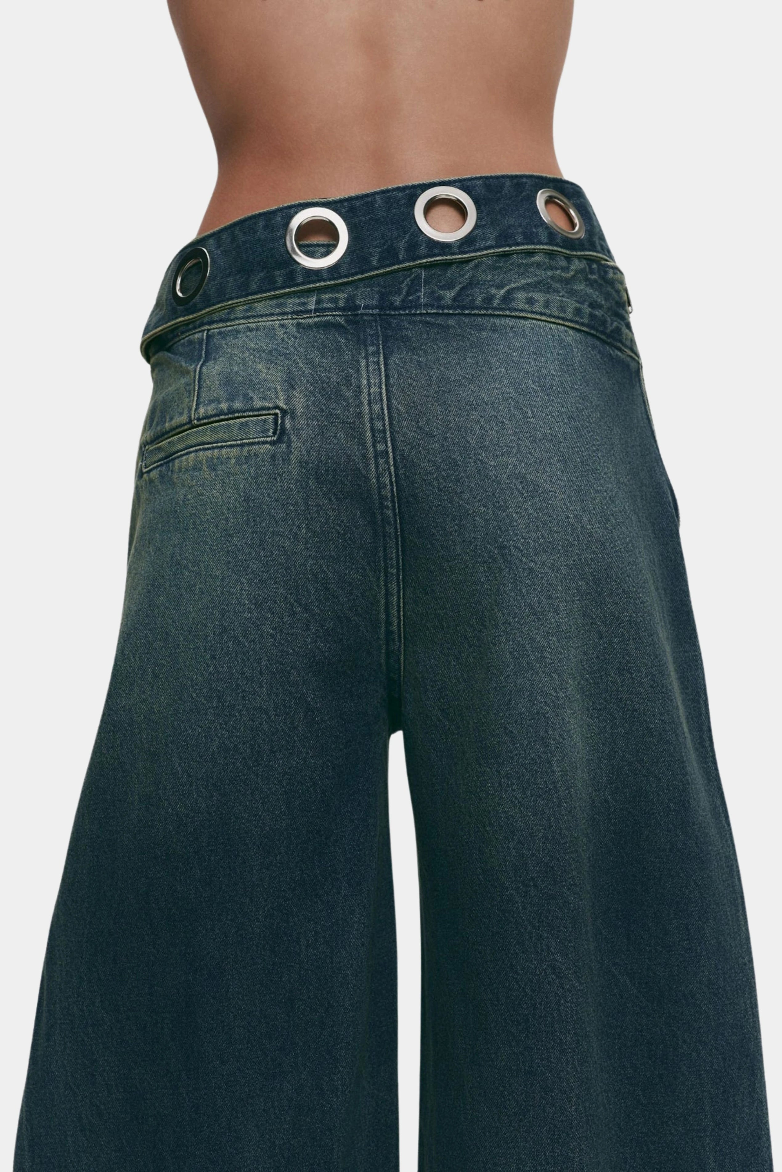 Detalhe do cós jeans azul escuro com ilhoses metálicos e acabamento assimétrico