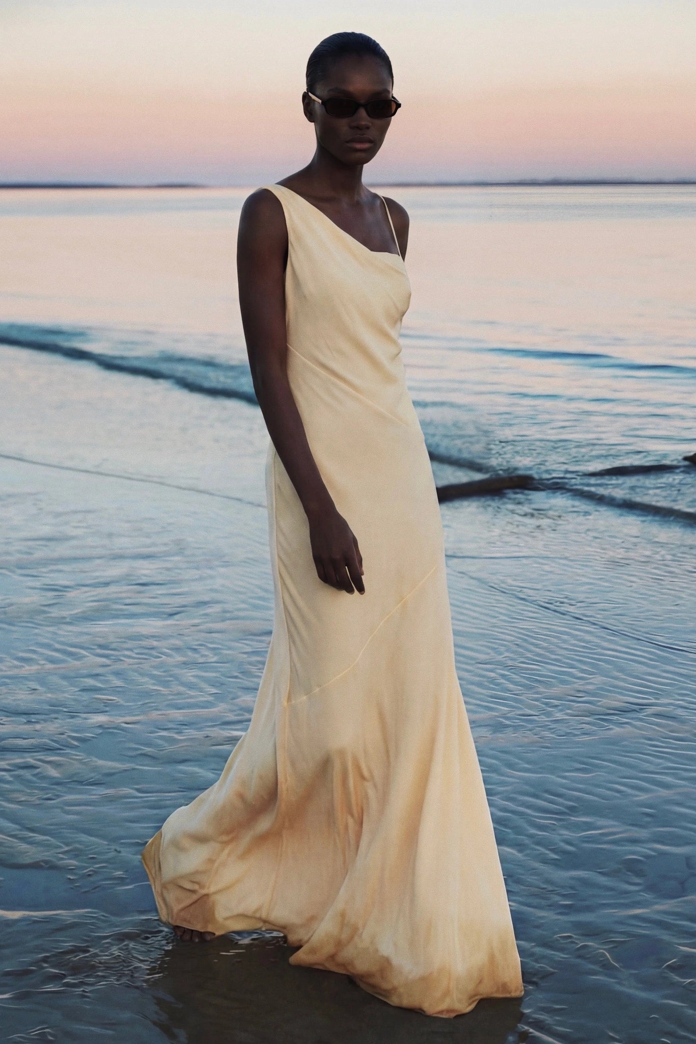 Modelo usando vestido longo amarelo claro assimétrico à beira-mar ao entardecer