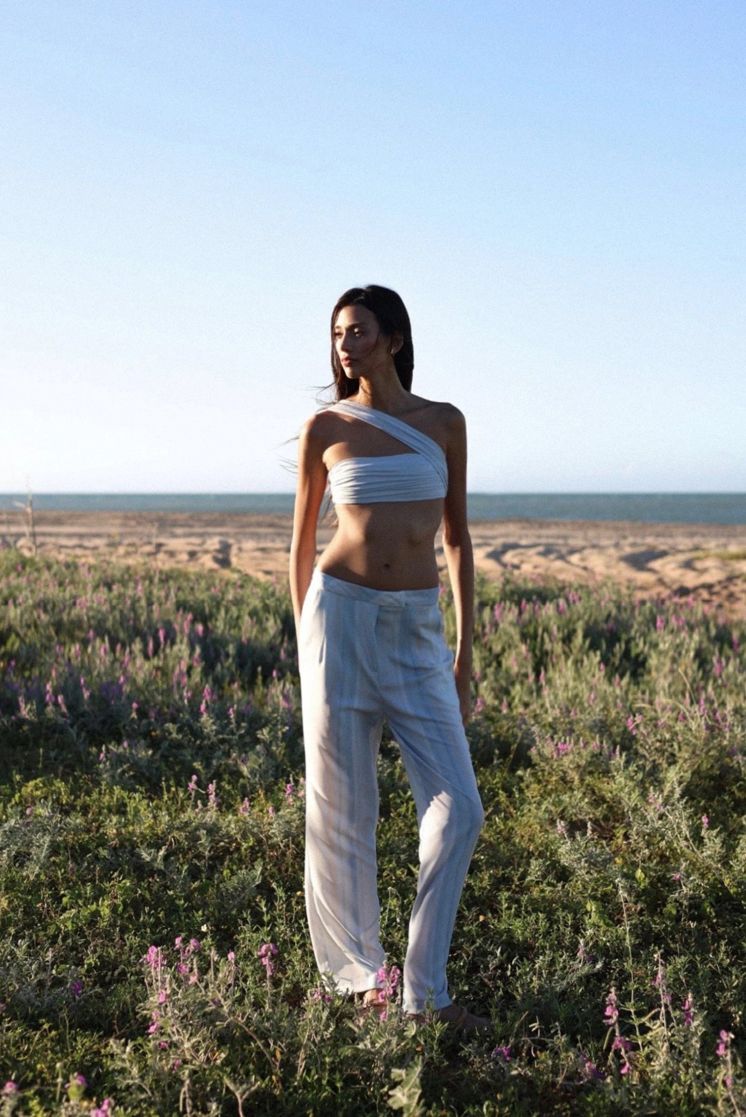 Look resort com top reto azul-claro de um ombro só e calça ampla em cenário de praia.