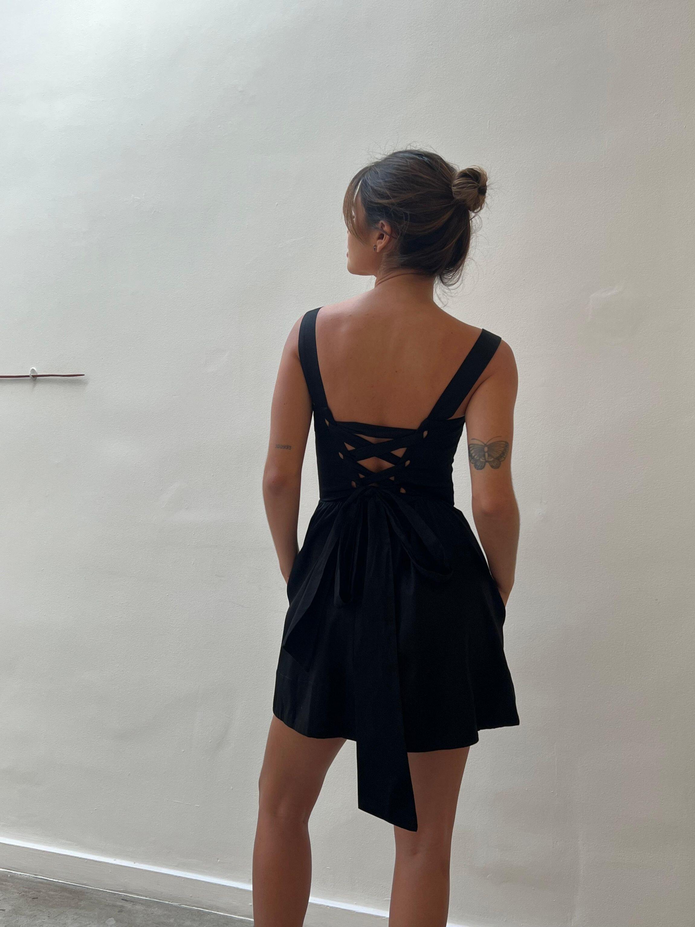 Modelo com vestido curto preto de costas com amarração cruzada e laço posterior